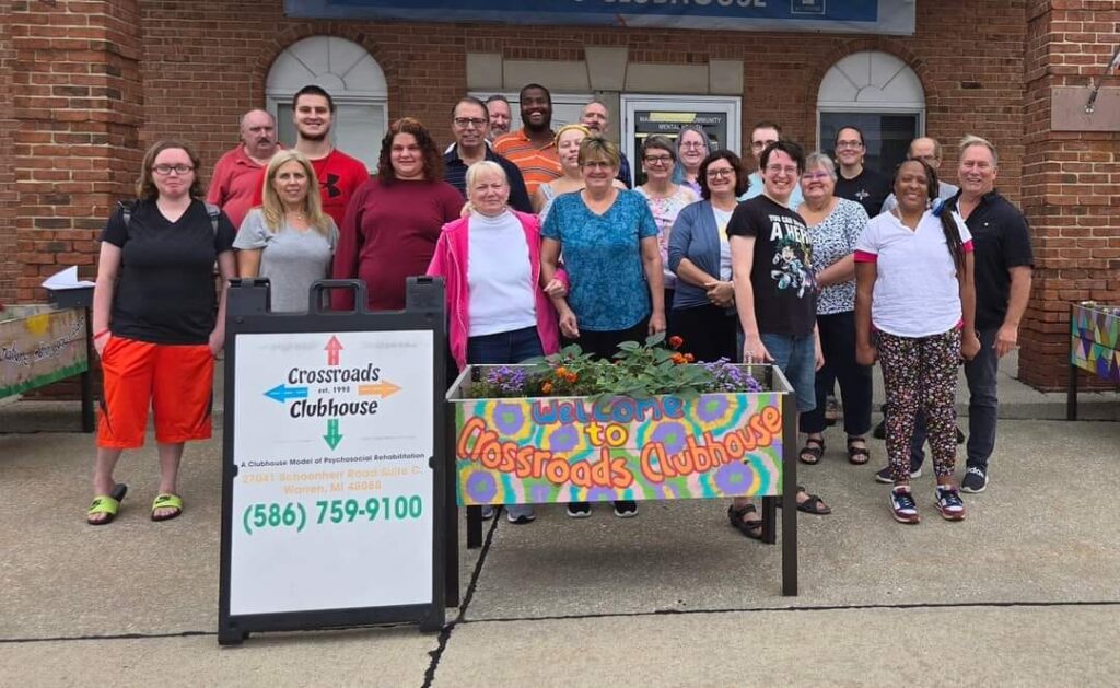 488879322_1216975283548556_1752413003970074085_n A group of people smiling standing in front of a building behind a sign that reads ‘Welcome to Crossroads Clubhouse.’