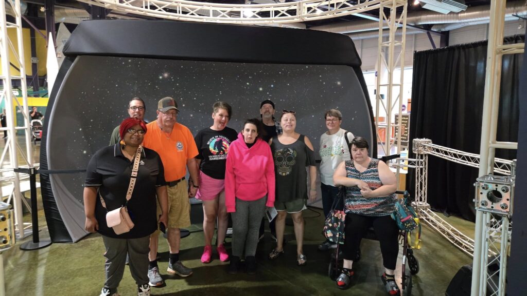 536280244_1326572742588809_4986377140272689989_n Group of people smiling in front of a galaxy backdrop at a planetarium.