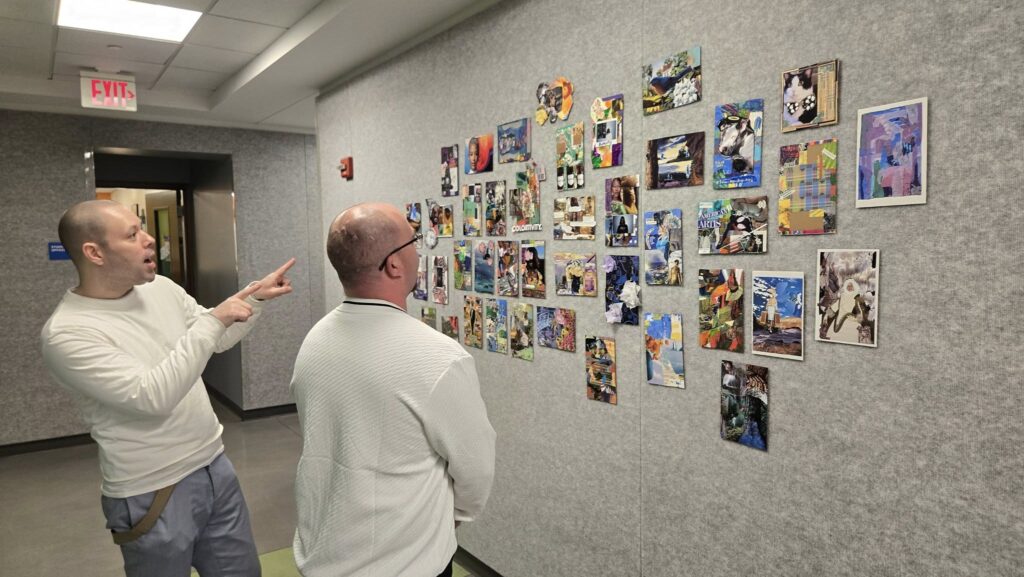 573073602_1385536366692446_4993855478417918208_n Two Caucasian men view collage art on display, with one man pointing at the artwork.