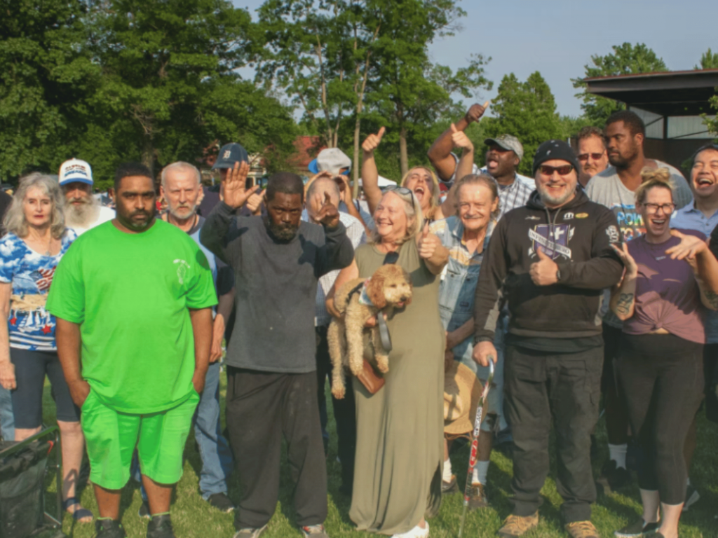 Liberties Inc. Photos A group of smiling individuals standing together outside on a sunny summer day, all giving a thumbs up, with trees visible in the background.