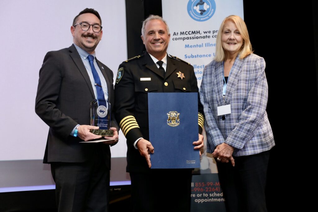 197A2787 (1) Three people stand on stage at an award event, with one holding a glass award and another in uniform holding a blue certificate folder in front of a community mental health banner.
