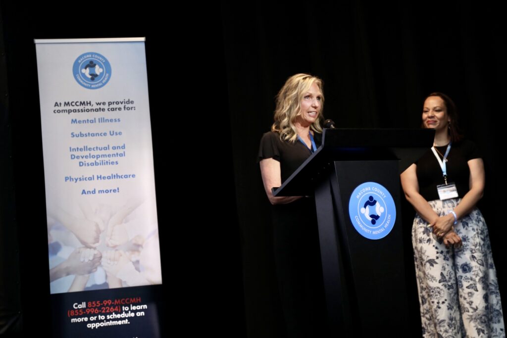 197A2802 Speaker at a podium on stage beside another person, with a community mental health services banner in the background.
