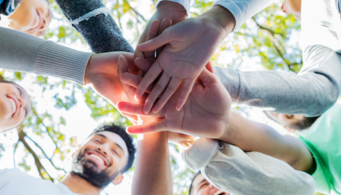 People standing outdoors form a circle and stack their hands together, viewed from below with trees and sunlight in the background.
