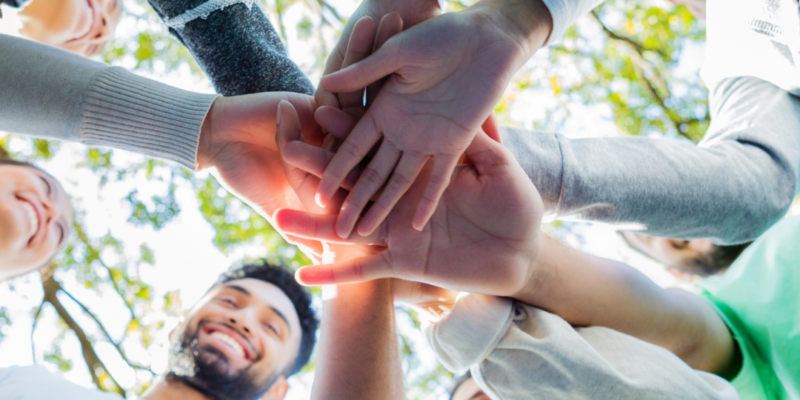 People standing outdoors form a circle and stack their hands together, viewed from below with trees and sunlight in the background.